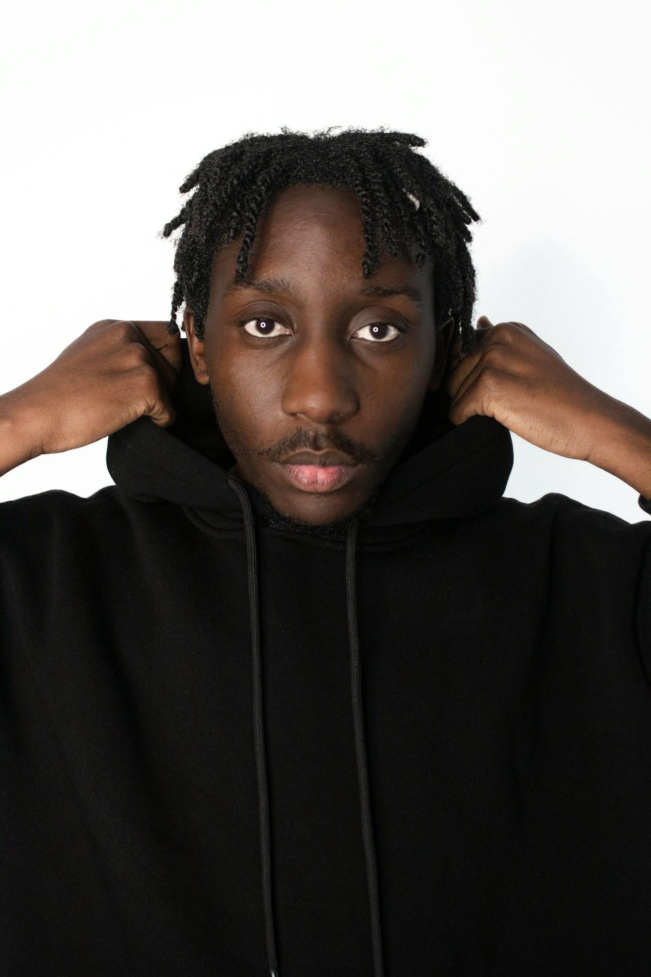 Man pulling up the hood of a black hoodie against a white wall