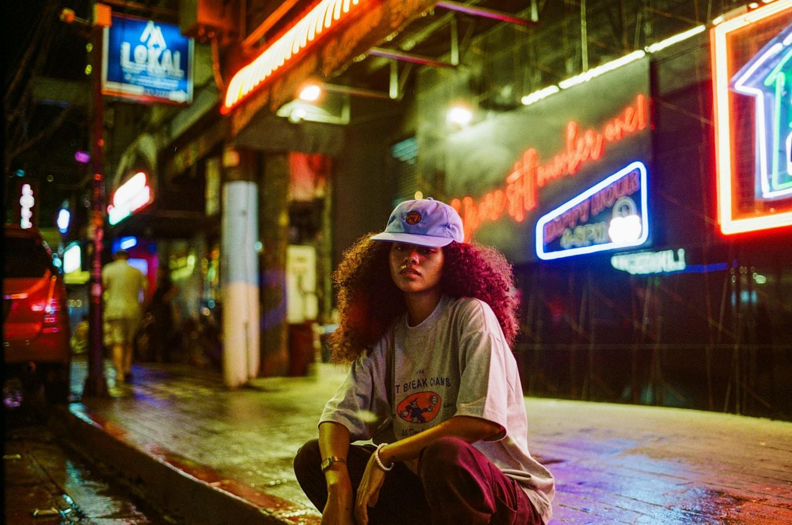 Woman crouched on a wet city sidewalk under neon signs at night